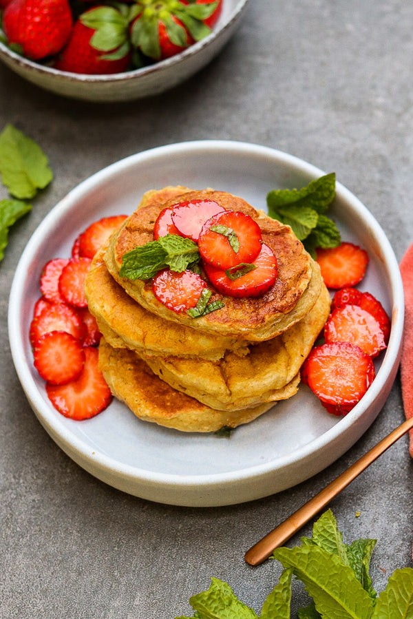 Ein Teller mit einem Stapel goldbrauner Banana-Pancakes, garniert mit frischen Erdbeerscheiben und Minzblättern. Daneben liegen weitere Erdbeeren und Minze, im Hintergrund eine Schale mit ganzen Erdbeeren.