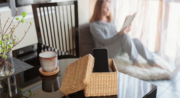 Smartphone in einer geflochtenen Box auf einem Tisch, während eine Frau im Hintergrund ein Buch liest – Symbol für digitale Auszeit.