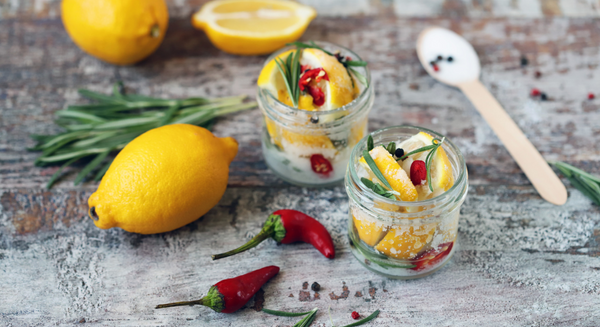 Fermentierte Zitronen in kleinen Gläsern mit Rosmarin, Chili und Pfefferkörnern, daneben frische Zitronen, Kräuter und Chilischoten auf rustikalem Holztisch.
