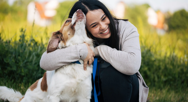 Lächelnde Frau kniet im Gras und umarmt liebevoll ihren Hund.