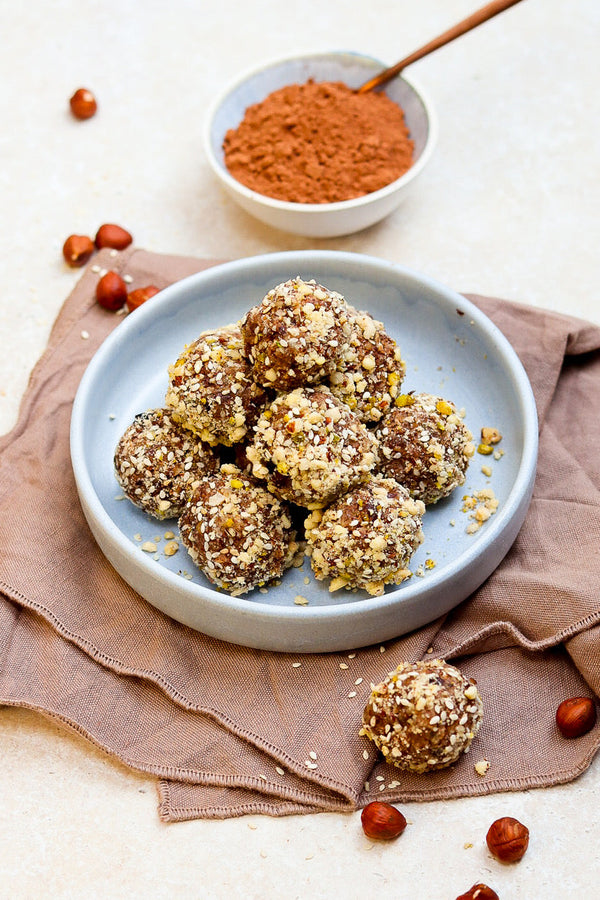 Ein grauer Teller mit selbstgemachten Energyballs, umhüllt von gehackten Nüssen, auf einem braunen Stofftuch. Im Hintergrund eine Schale mit Kakaopulver und verstreute Haselnüsse – ein gesunder Snack voller Energie.