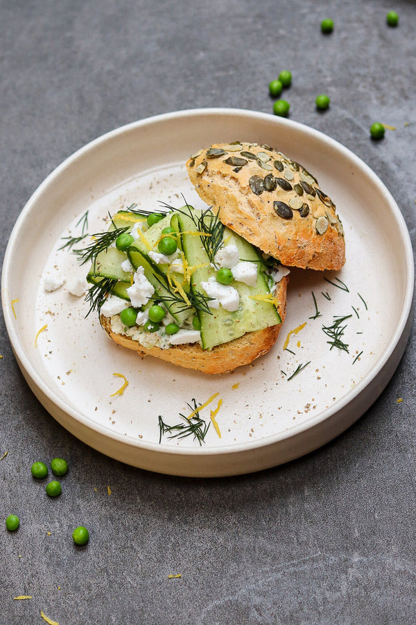Vollkornbrötchen mit Kürbiskernen, belegt mit grünem Erbsen-Aufstrich, Gurkenscheiben, Feta, frischem Dill und Zitronenzeste, serviert auf einem hellen Teller.
