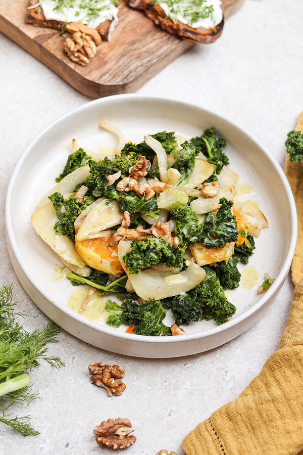 Grünkohl-Fenchel-Salat mit Orangenfilets, Walnüssen und cremigem Dressing auf einem weißen Teller, im Hintergrund Vollkornbrot mit Frischkäse und Kresse.