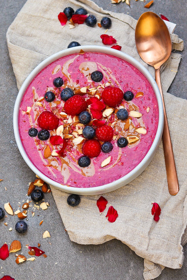 Pinkfarbene Smoothie-Bowl mit Himbeeren, Blaubeeren, gehackten Mandeln, Mandelmus und essbaren Blüten – sommerlich angerichtet auf einem Leinentuch mit Kupferlöffel.