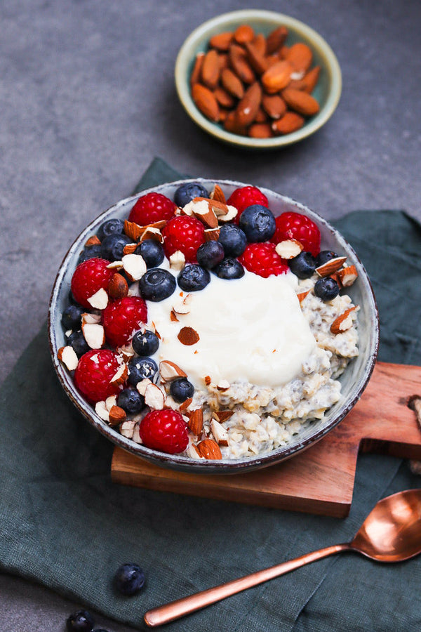 Schale mit warmem Porridge, getoppt mit frischen Himbeeren, Heidelbeeren, Kokosjoghurt und gehackten Mandeln, daneben eine Schale mit Mandeln.